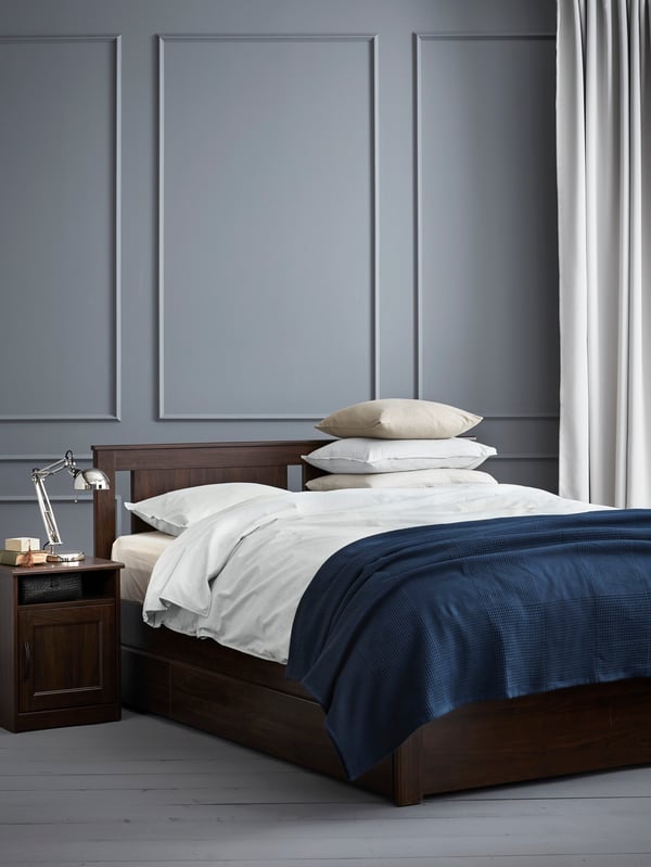 Bedroom with brown wooden bed frame, bedside table, duvet cover and pillowcases, bedspread, curtains, cushion covers, lamp.
