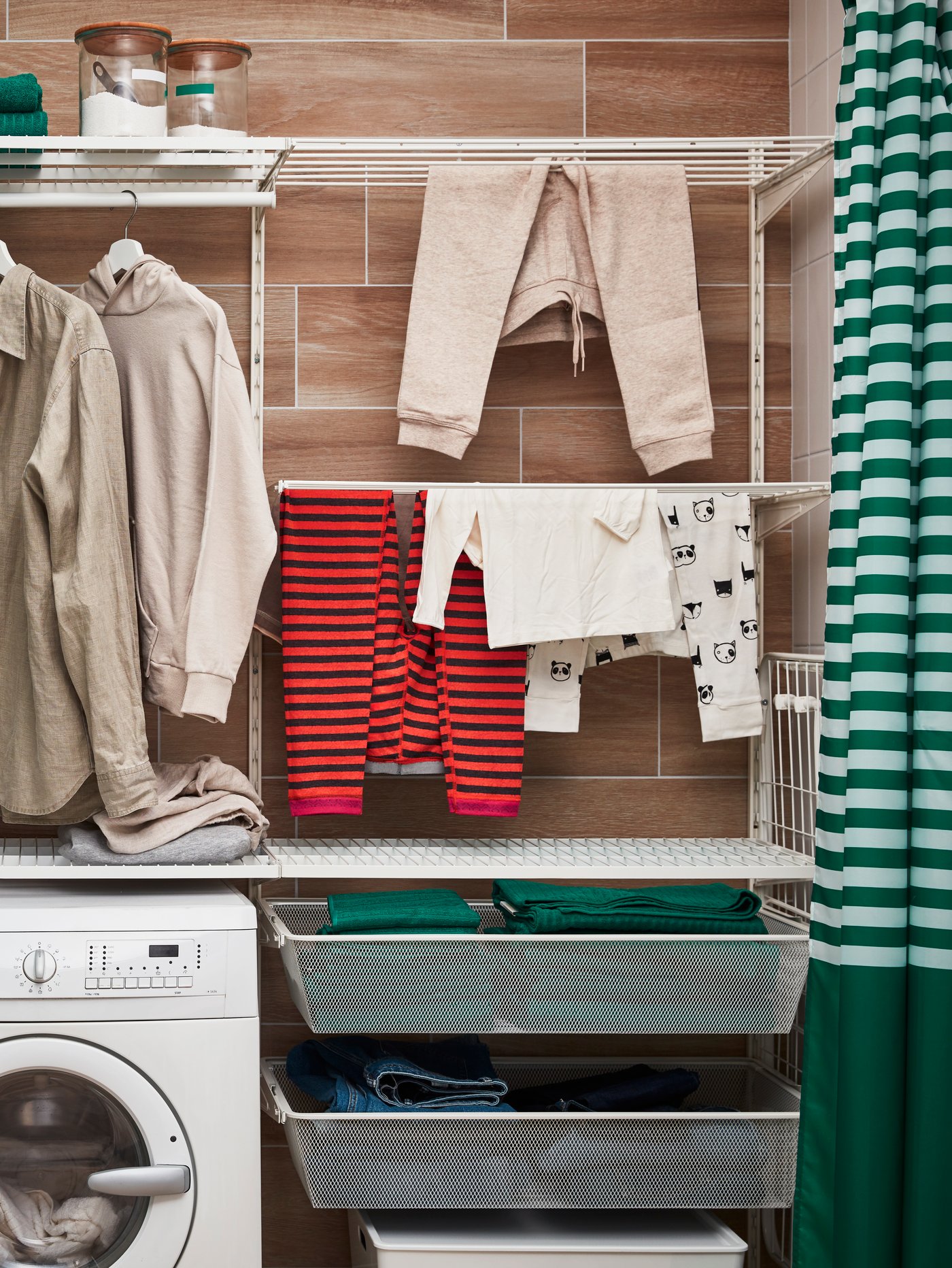 Vista frontal de colgadores, canastos y repisas BOAXEL que arman un espacio de lavandería con la lavadora incluida y ropa colgada. El muro del fondo es de cerámica con un patrón café claro, y frente a las repisas hay dos cortinas a rallas blanco con verde que se pueden cerrar para ocultarlo.