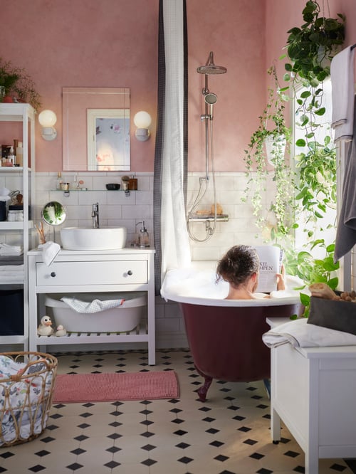 Ein Badezimmer im traditionellen Stil mit weißen Fliesen, rosa Wänden und grünen Pflanzen. Eine Person mit langen Haaren sitzt in der Badewanne und liest eine Zeitschrift. 