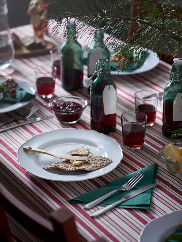 A decorated holiday table set with a beige-and-red VINTER 2021 tablecloth and one VINTER 2021 bottle by each UPPLAGA plate.