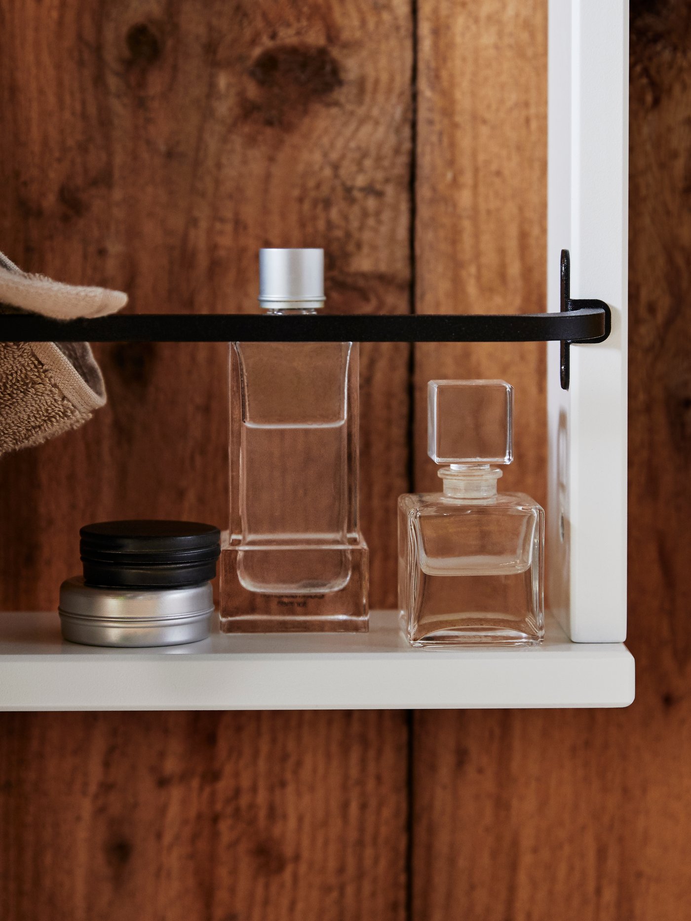 Ein weißes HEMNES Wandregal mit zwei Parfümflaschen und zwei Metallbehältern. Über der schwarzen Stange hängt ein braunes Handtuch.