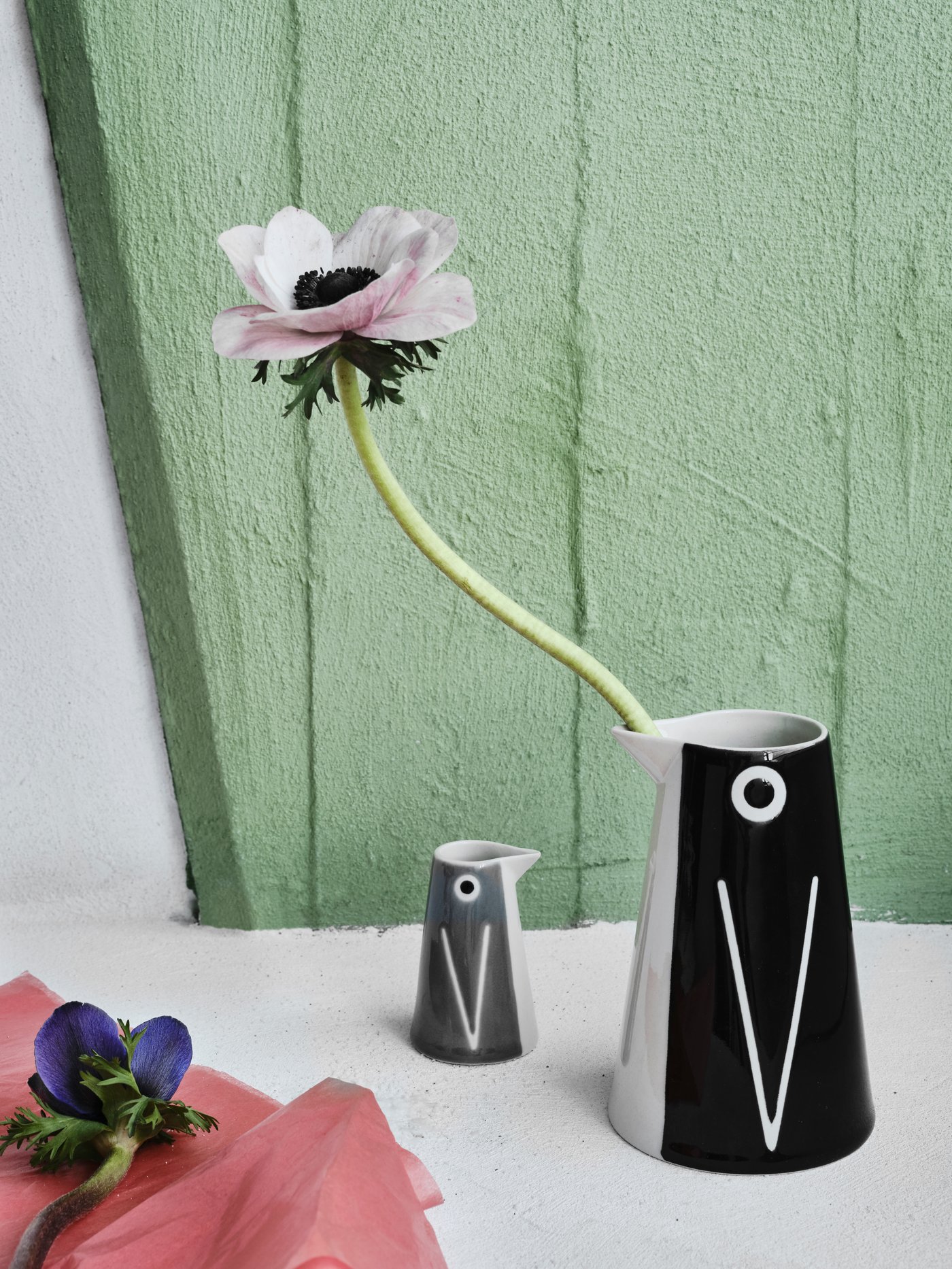 Vaze/carafe LÄTTSÅLD de mici și mari dimensiuni în formă de pinguin pe o masă, iar în vaza mai mare se află o floare.