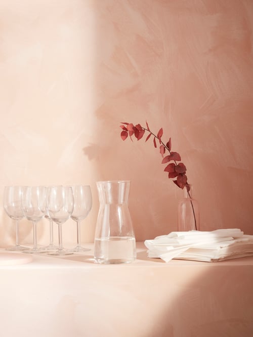 Eine Stufe mit mehreren SVALKA Weingläsern, einer KARAFF Karaffe mit Wasser, einer Vase mit einer roten Blume und Papierservietten.