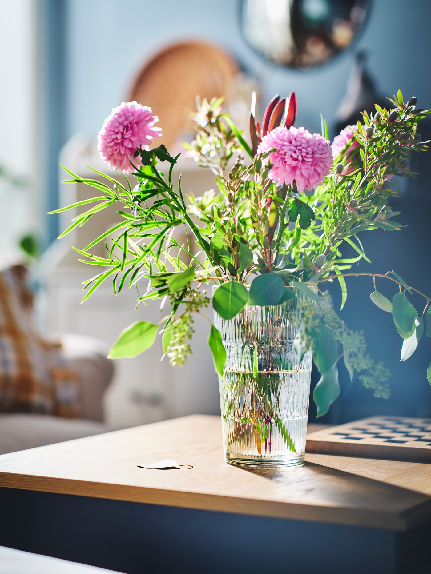 In einem Wohnzimmer steht eine SMÄLLSPIREA Vase aus Glas mit Blumen im Inneren auf einem SKRUVBY Couchtisch in Schwarzblau.