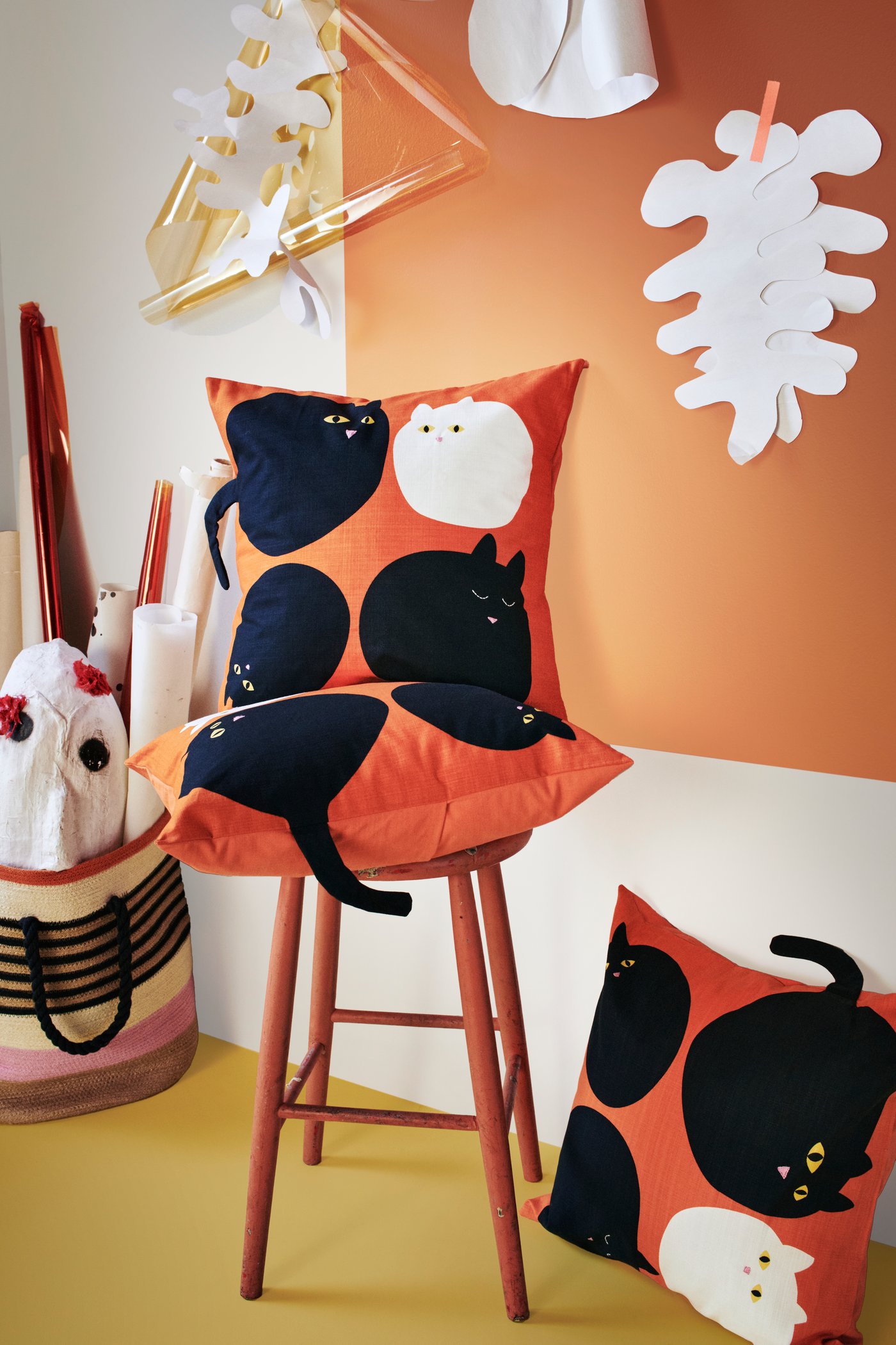 Dos fundas de cojín MÄVINN naranjas con un estampado de gatos colocadas en un taburete alto, y uno de ellos en el piso.