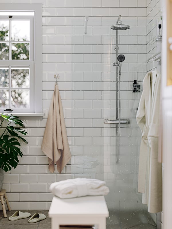 A VOXNAN shower in the corner of a white tiled bathroom beside a towel on a hook. A clear shower screen is in front of it.