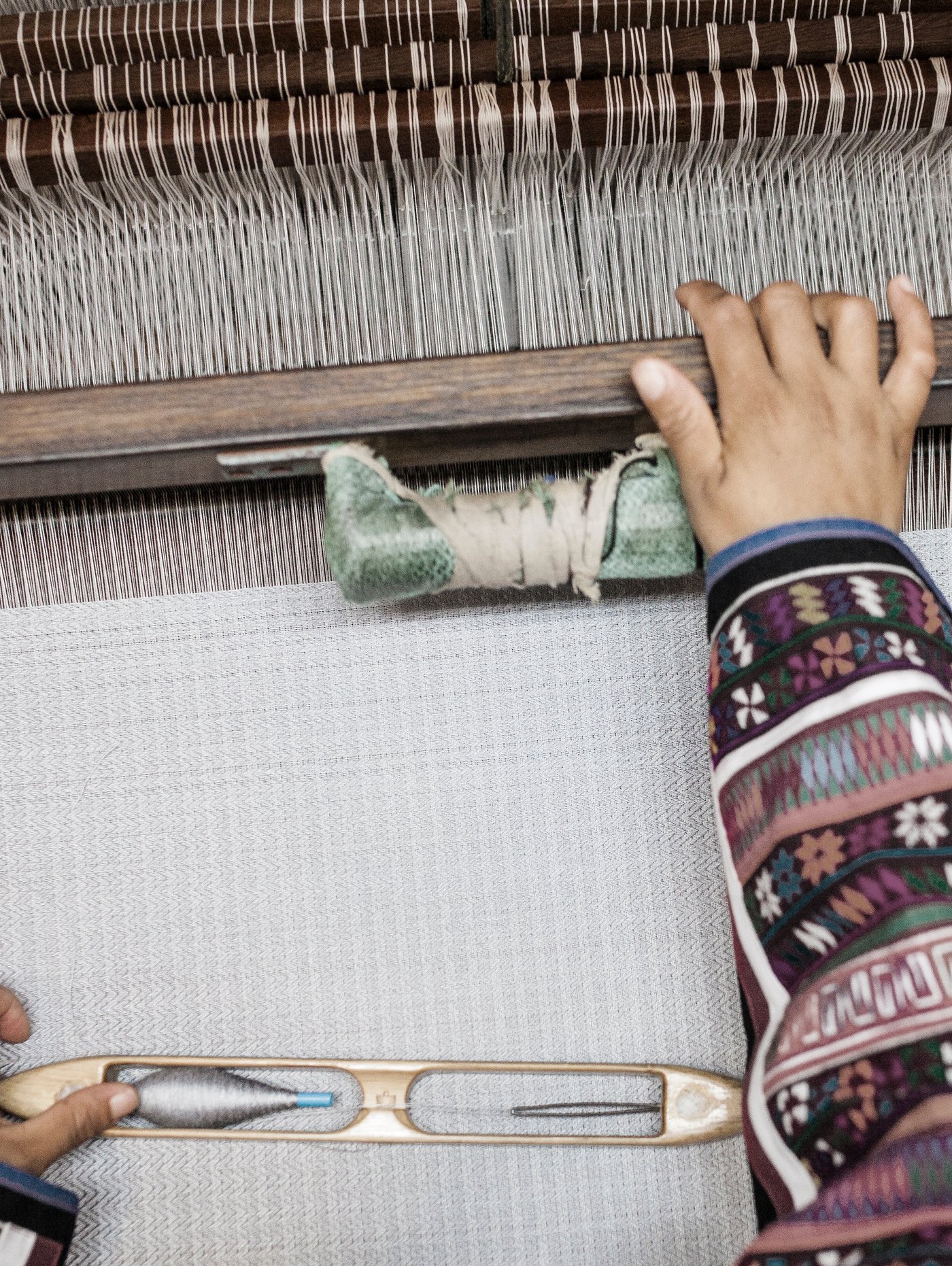 The hands of a weaver wearing a traditionally patterned top in the process of weaving a white rug on a loom.