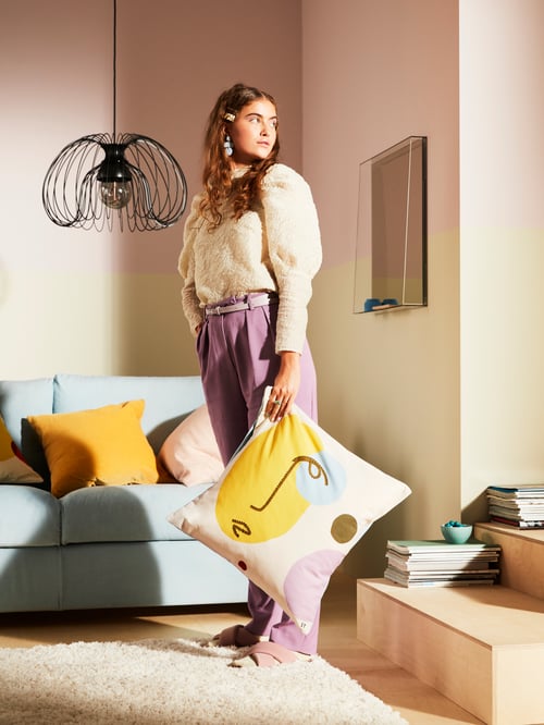 A black pendant lamp shade hangs behind a person holding a square cushion in one hand. Behind the person is also a sofa.