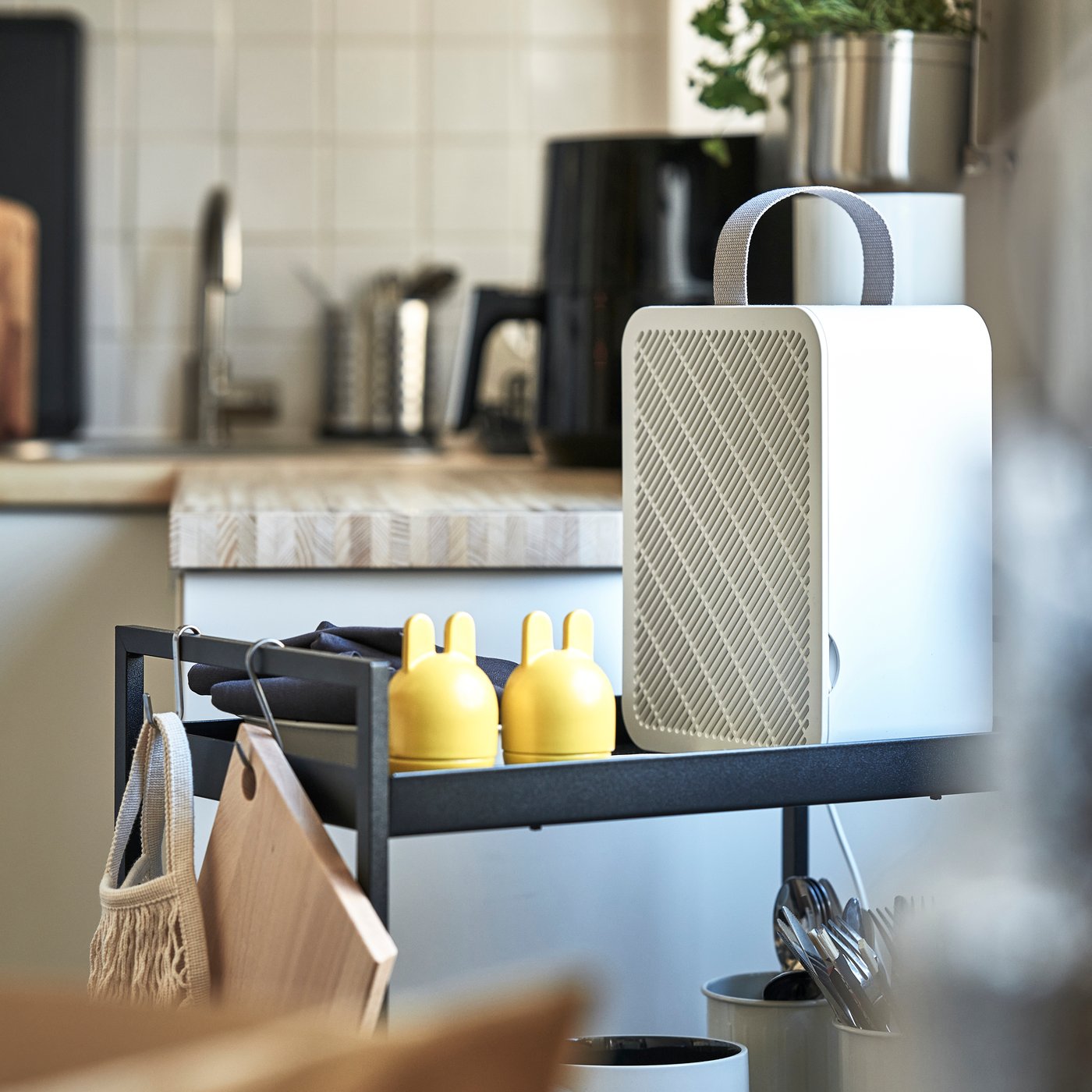 Rektangulär vit luftrenare med handtag står på en hylla i ett kök, bredvid två små gula prydnadsföremål.