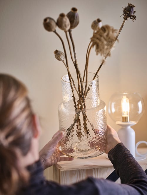 Woman holding clear patterned vase made of mouth-blown leftover glass holding dried flowers, over cabinet with table lamp.