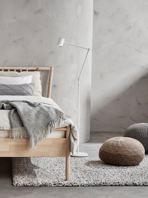 Dormitor minimalist cu pereți gri, cadru de pat din lemn, lampă albă de podea, aruncare gri, covor înalt și pouff-uri.