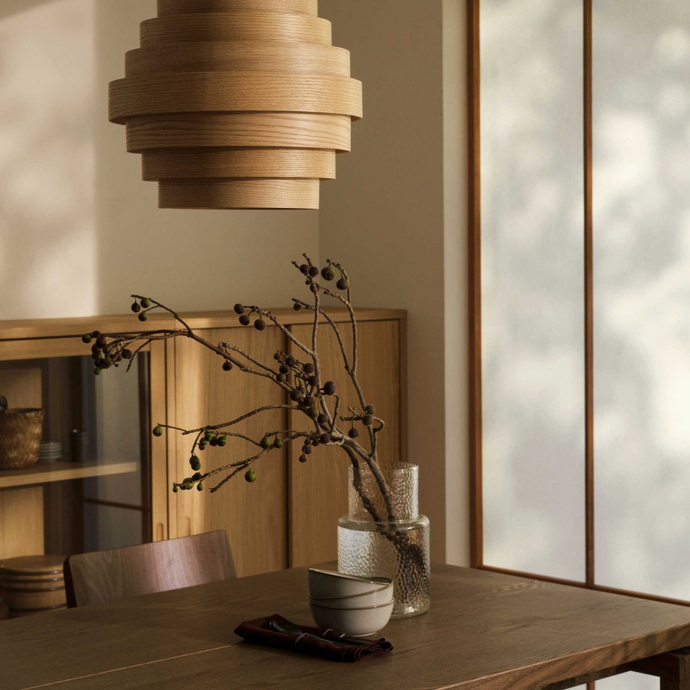 Minimalistiskt rum i japandi-stil med träbord, dekorativ gren i vas och modern DYKARKLOCKA trälampa som hänger från taket.

