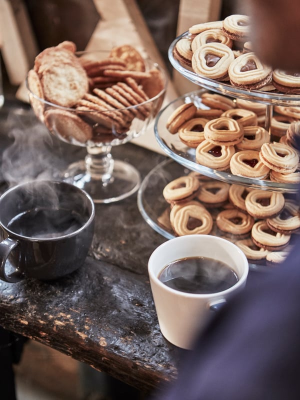 Serveringsfad i 3 etager fyldt med hjerteformede småkager, 2 krus med varm kaffe og en skål med runde småkager.