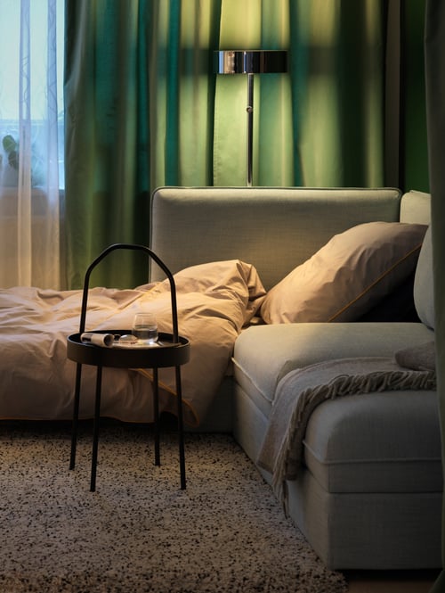 Living room with sofa, black side table, glass, black floor lamp, beige throw, green room darkening curtains, high pile rug.