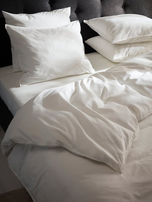 Bedroom close-up of a white ÄNGSLILJA duvet cover with 2 pillowcases placed on top of a sateen NATTJASMIN fitted sheet.