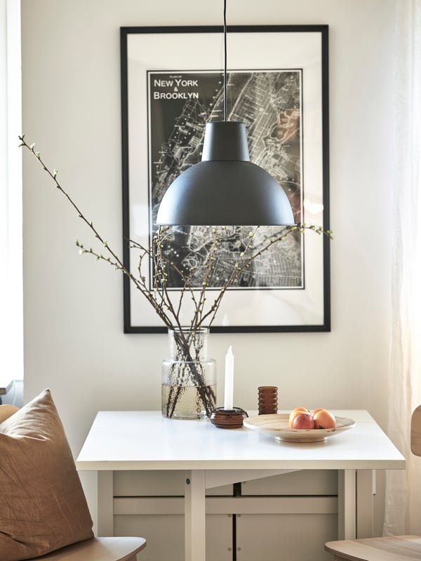 En flexibel matplats med ett vitt slagbord och två stolar i ljust trä. Ovanför matbordet hänger en svart lampa och bakom syns en stor tavla med stadsobjekt.