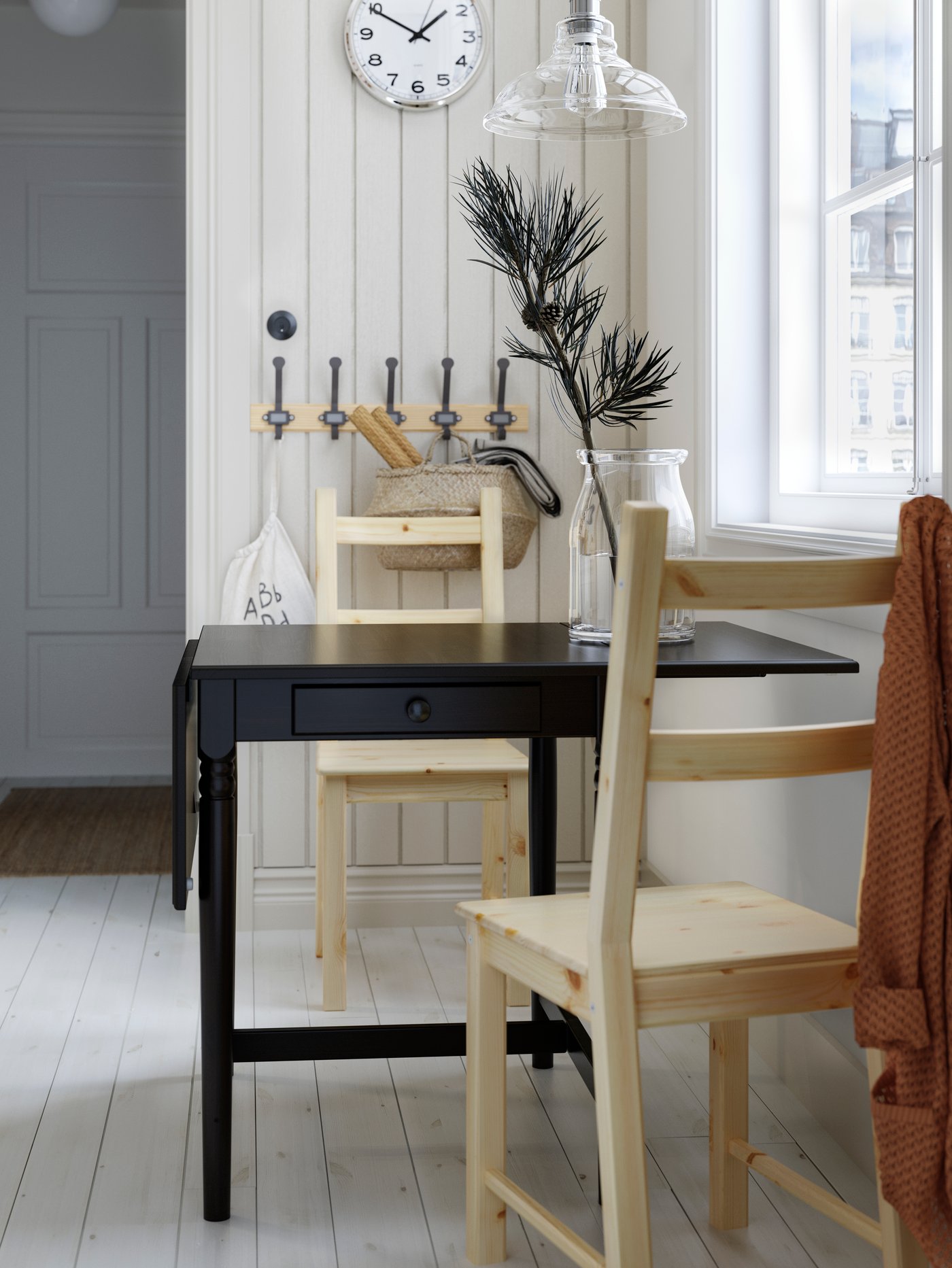 A black drop-leaf table with drawer, two pine chairs, a wooden rack with hooks, a wall clock and a glass pendant lamp.