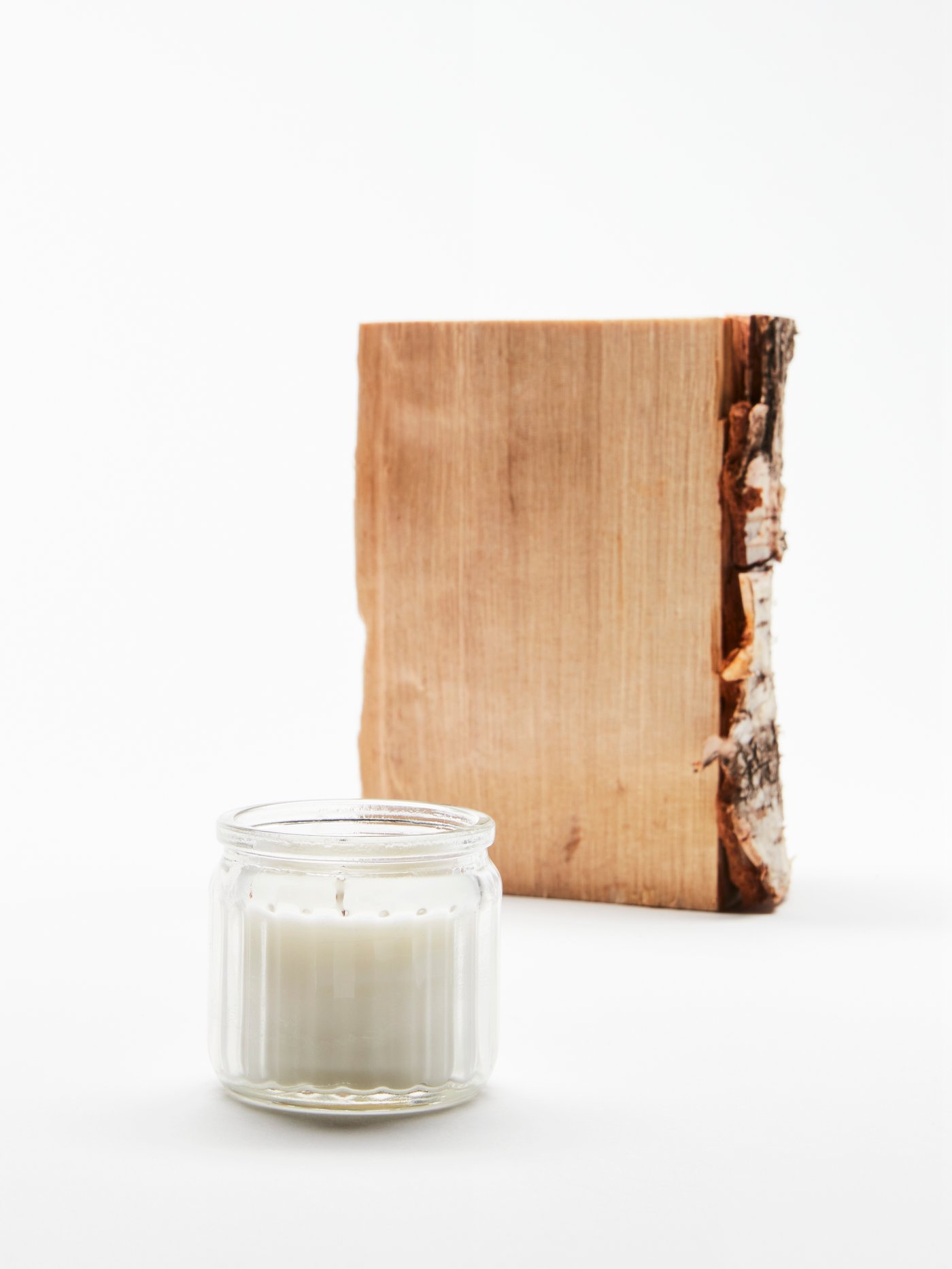 An ADLAD scented candle in glass is in the foreground of a white studio space. Behind it stands one birch wood log.