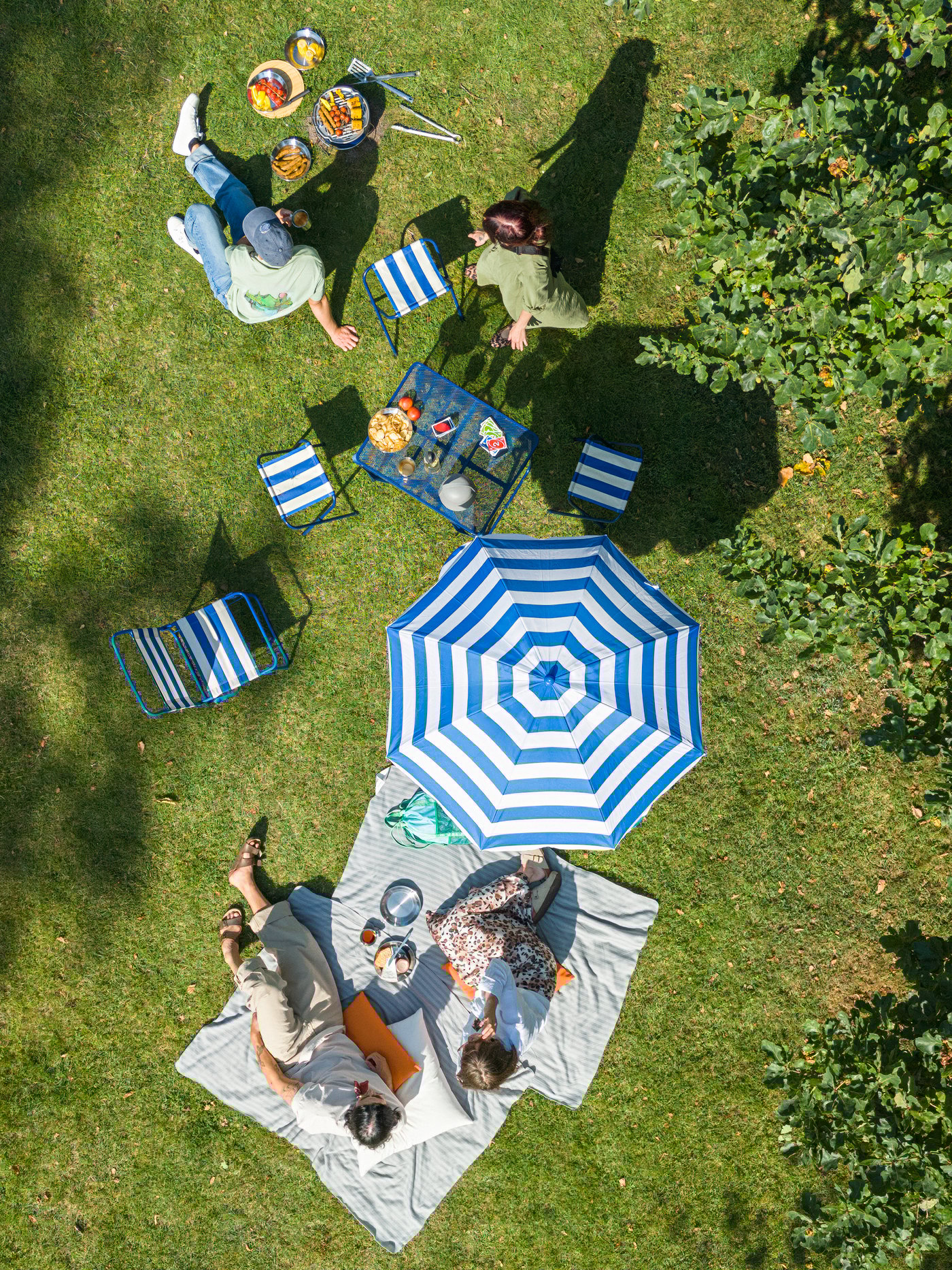 Friends have a barbecue in the park, relaxing on the grass with a STRANDÖN table, chairs, stools, and parasol.