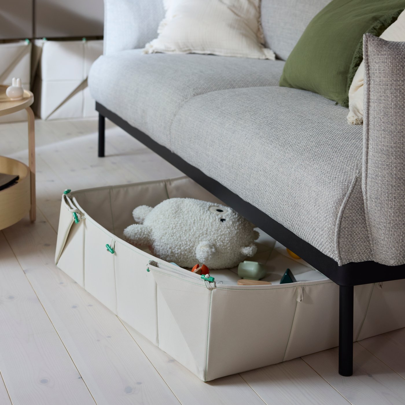 En beige GREJSIMOJS måtte/sammenfoldelig opbevaringskasse stikker ud fra under sofaen i en moderne stue. Der er bløde tøjdyr i kassen.