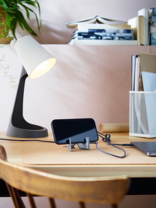 Un spațiu de lucru care dispune de un smartphone într-un suport de telefon mobil pe un suport de birou, un cablu USB de la A la USB C și o lampă de lucru.