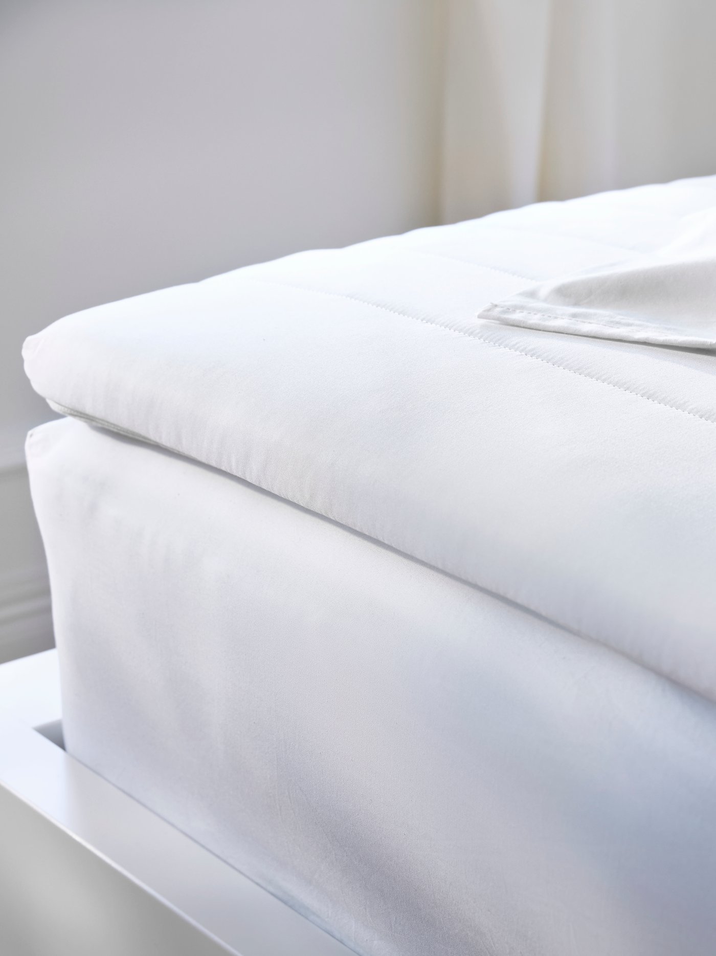 Close-up of corner of a bedframe, mattress and mattress pad with sheets in a pile on top, all in crisp white.