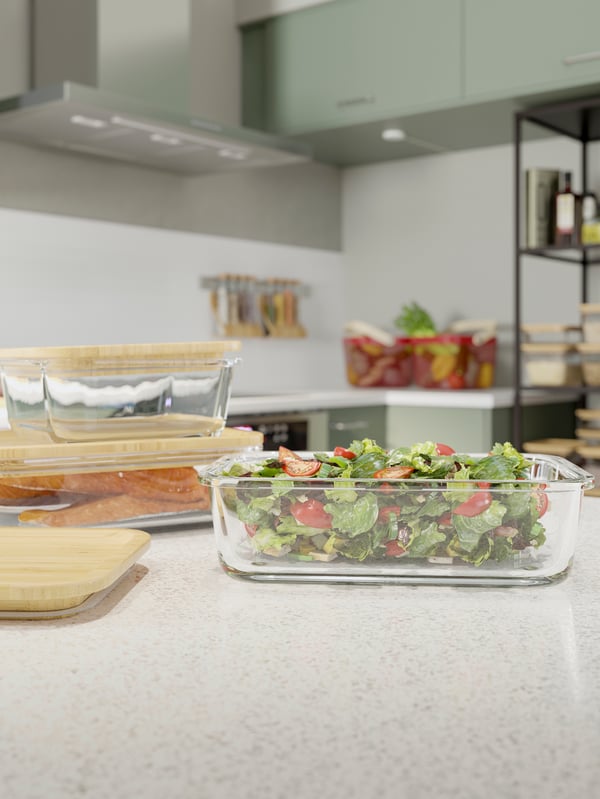 A close-up of two IKEA 365+ glass food containers with bamboo lids and one without storing salad on a worktop.
