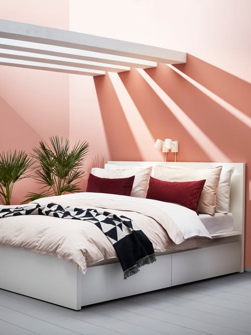 Bedroom with pink walls, wooden beams above white bed frame with high headboard, bed linens, pillows, throw, and wall lamps.