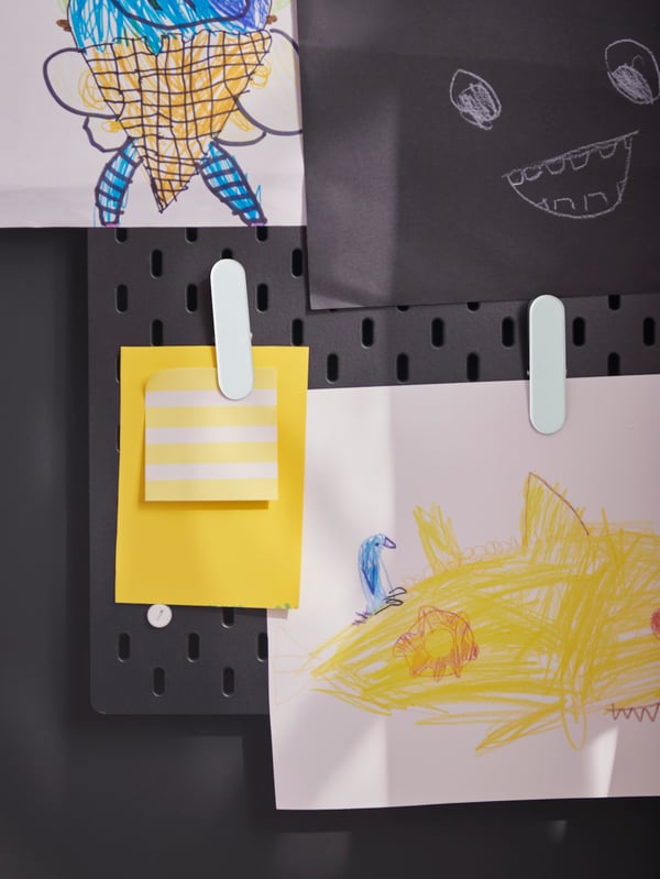 An einer schwarze SKÅDIS Lochplatte, die an einer schwarzen Wand hängt, sind farbiges Papier und eine Kinderzeichnung mit weißen SKÅDIS Klemmen angebracht.