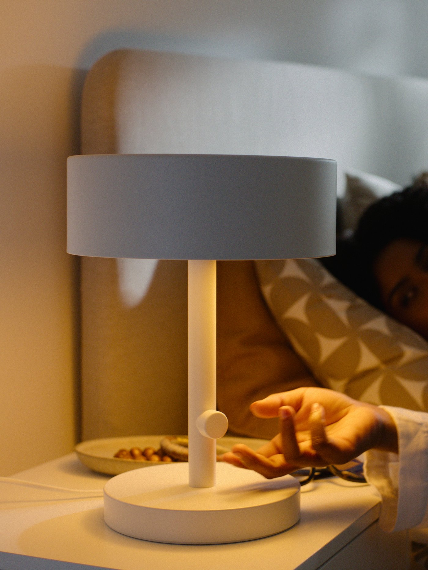 En vit NYMÅNE bordslampa på ett sängbord. En hand sträcker sig för att justera ljusstyrkan.
