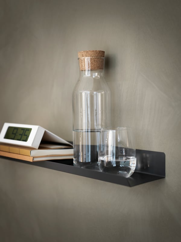 Minimalistisk svart MALMBÄCK vägghylla med glasflaska, tomt glas, två böcker och en digital klocka som visar 9:35.