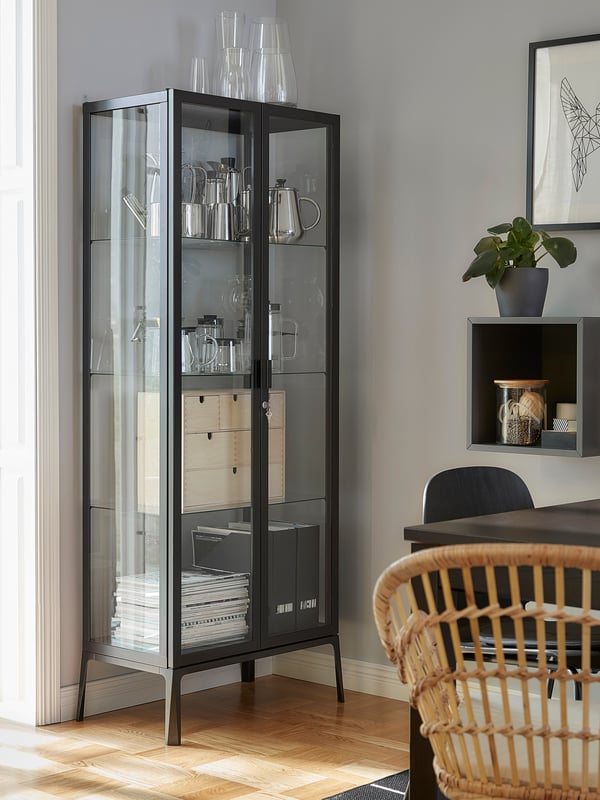 Armario MILSBO con portas de cristal a carón dunha porta de balcón. Dentro hai artigos decorativos, revistas e ficheiros con revistas.
