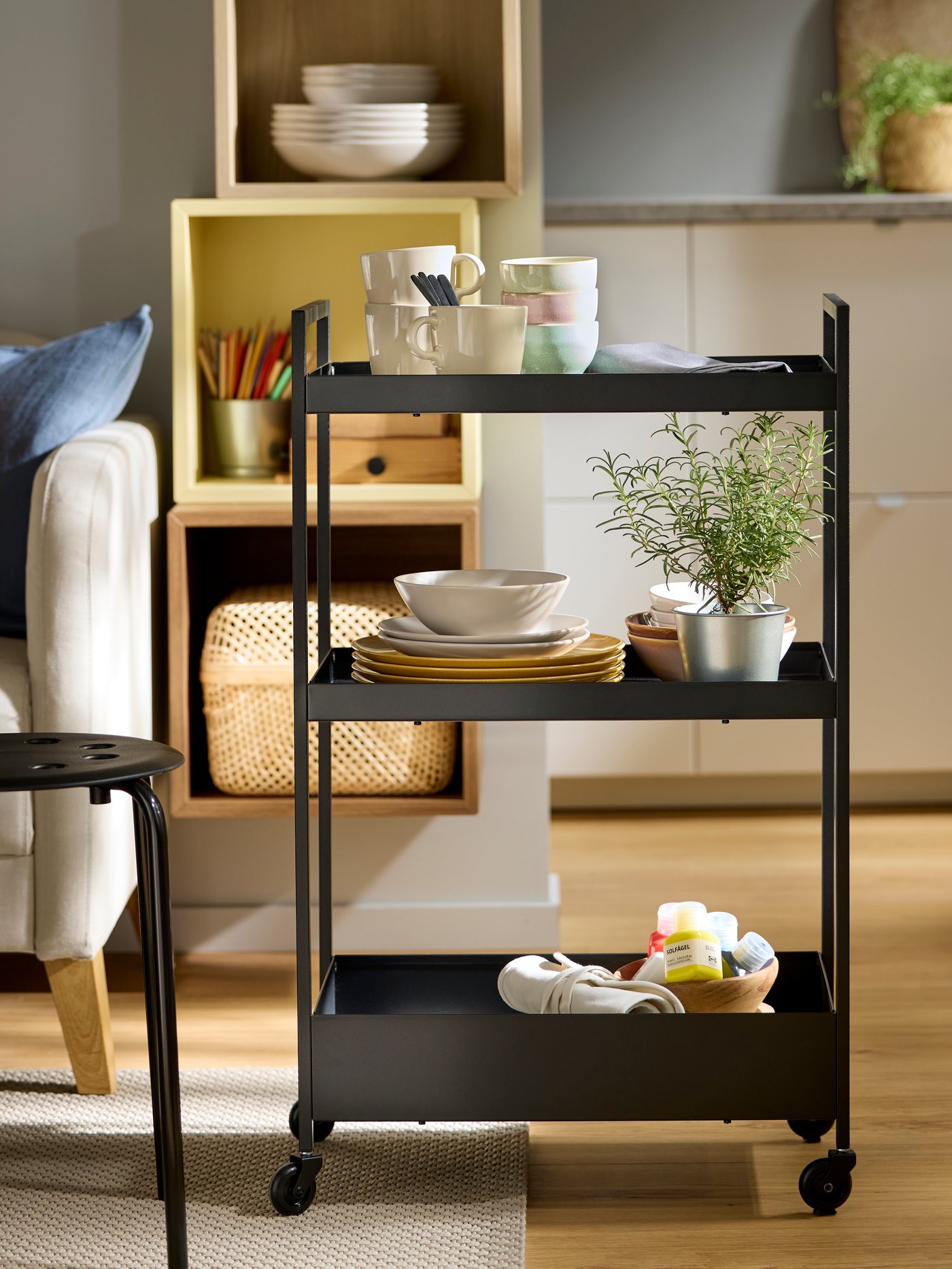 A black NISSAFORS trolley with three shelves filled with FÄRGKLAR plates and bowls in pastel colours, next to a MARIOUS stool.