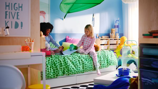 A children’s room with blue wall, two girls are playing on SLÄKT bed with green NATTHÄGER duvet cover and colourful cushions.