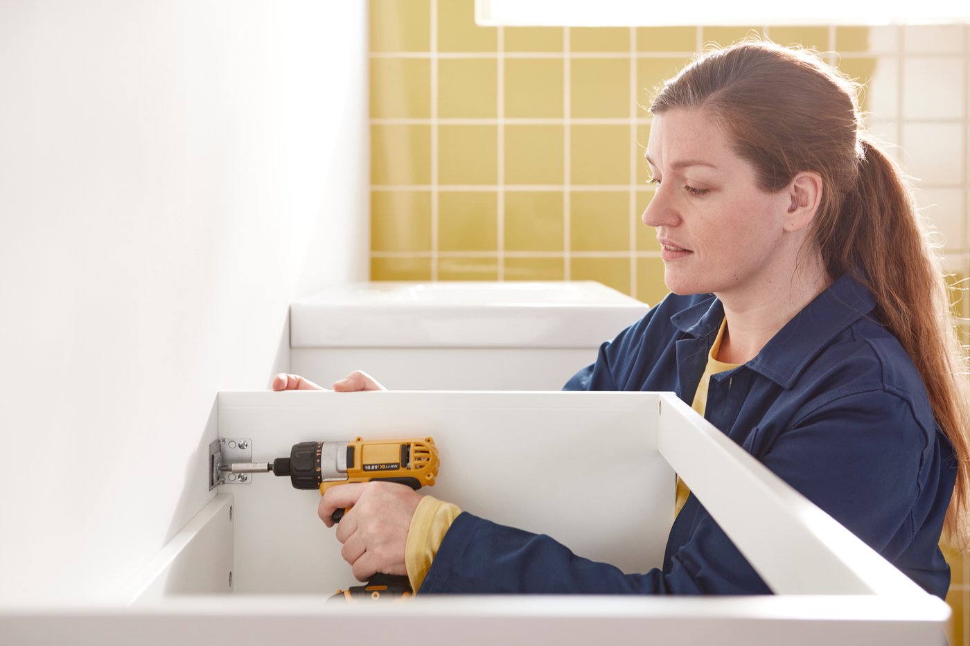 Une femme en bleu de travail utilise une perceuse pour fixer une armoire au mur.