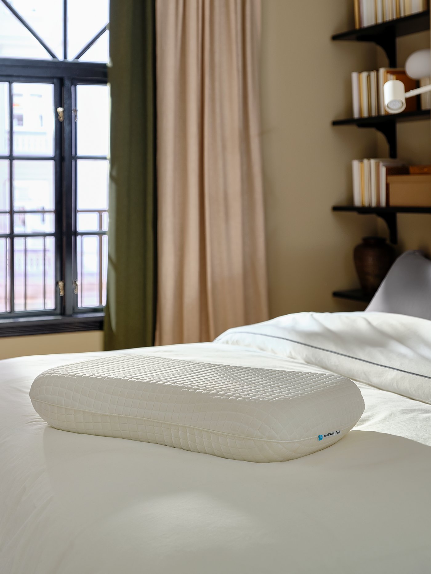 Un oreiller ergonomique sur un lit blanc près d’une fenêtre.