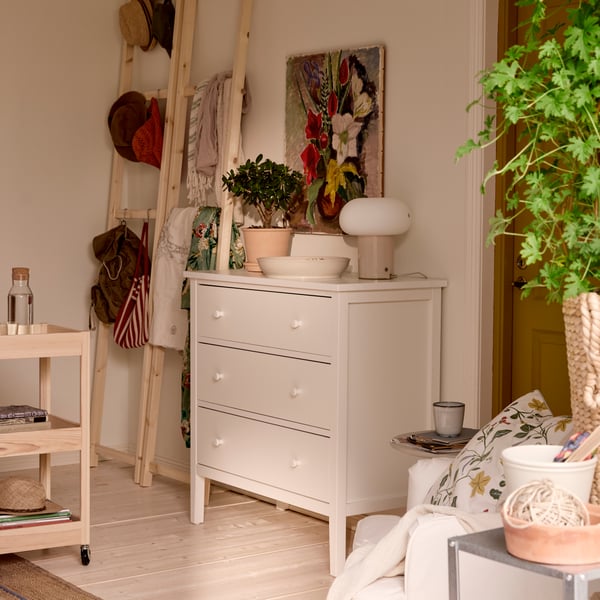 A white GULLABERG chest of drawers with pots and plants on top, a picture above it and wooden ladders in the back for storage.