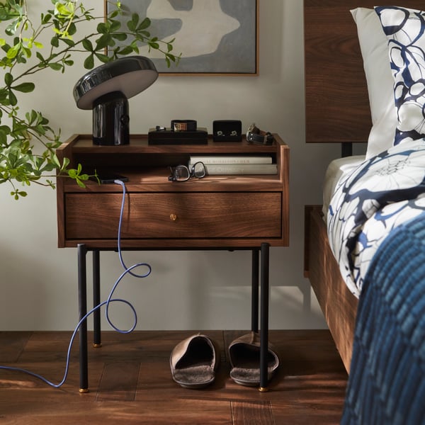 A bedroom featuring a brown RÅDMANSÖ bedside table with a walnut effect on legs, beside a bed and a potted plant.