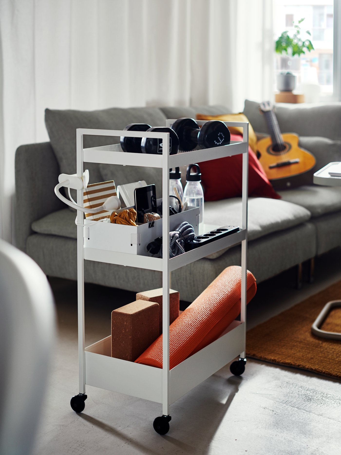 Biały wózek kuchenny NISSAFORS wypełniony matą sportową, klockami do yogi, i hantlami. Na drugim planie stoi szara sofa z gitarą.