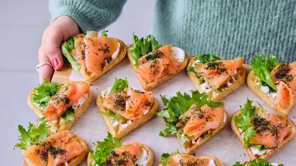 A person in a blue sweater is holding a wooden board and it has heart-shaped waffles with salmon, cream cheese and lettuce.