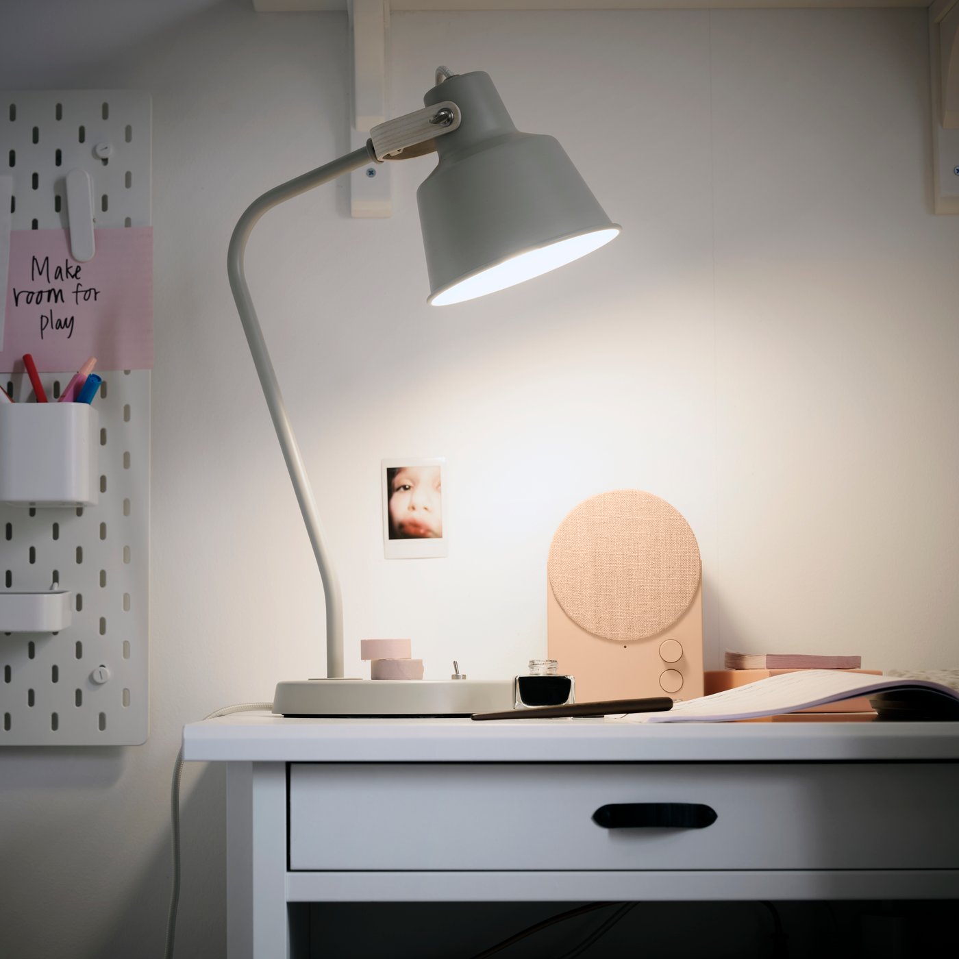 A study area features a beige SJÖMAKT desk lamp made from ash, illuminating a small table beside a photo and stationery.