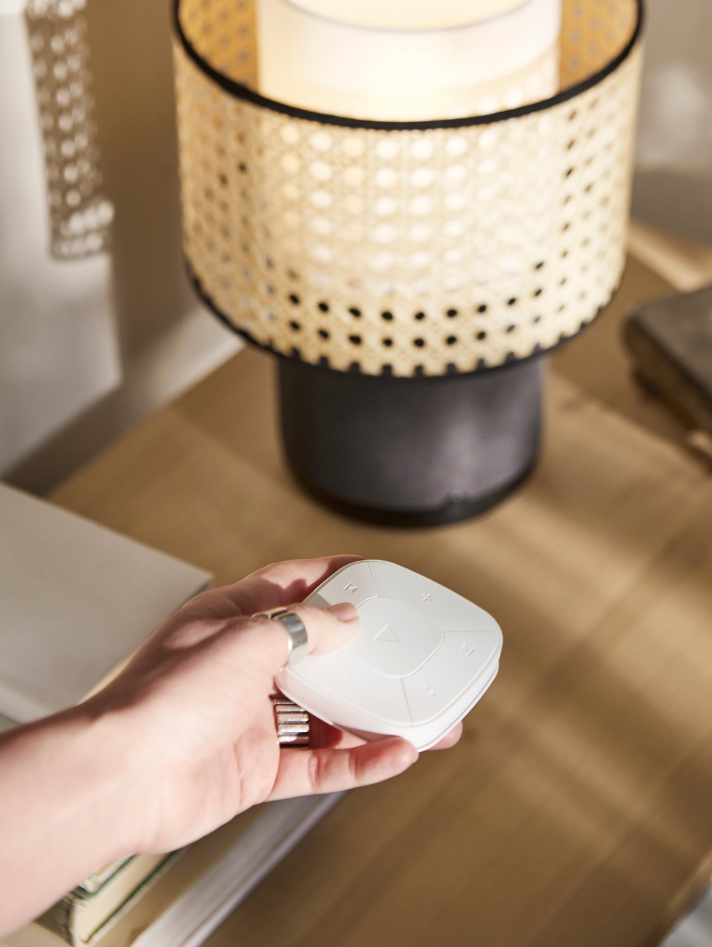 يد تحمل جهاز تحكم عن بُعد في الصوت بجوار غطاء مصباح خيزران SYMFONISK على مصباح سماعة صوت أسود.