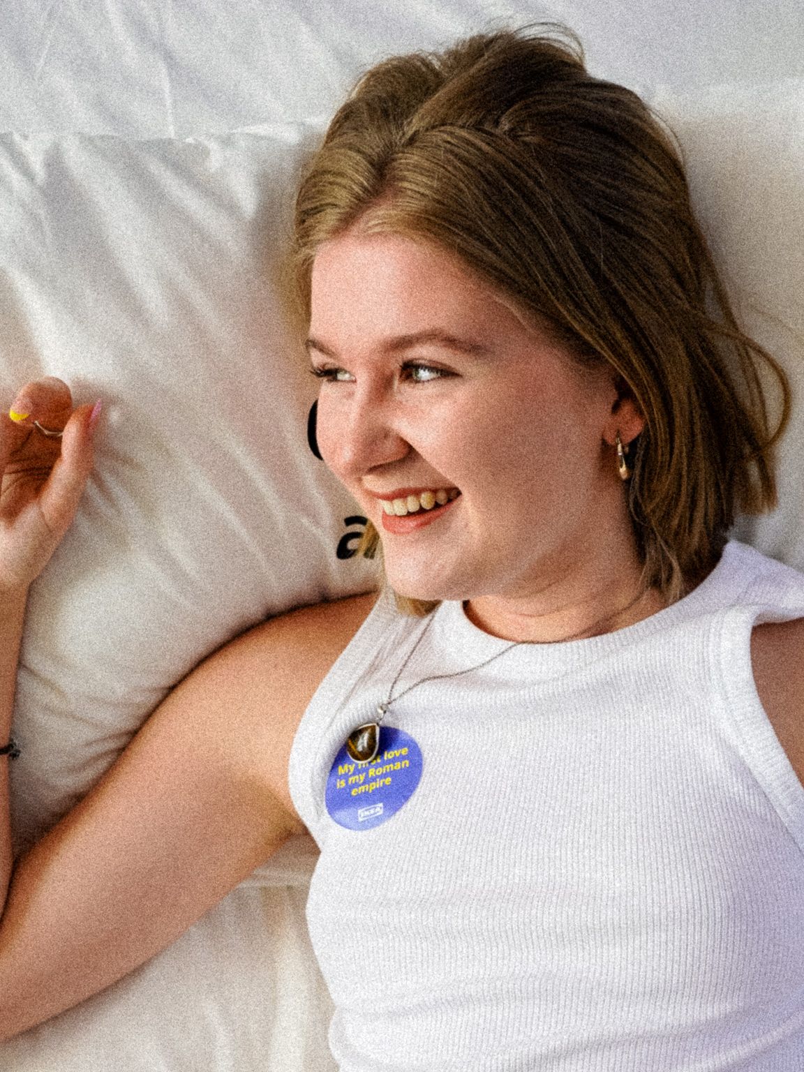 Izzy leži na postelji, oblečena v belo majico. Smehlja se in obraz obrača v desno, ob njem pa drži desnico.