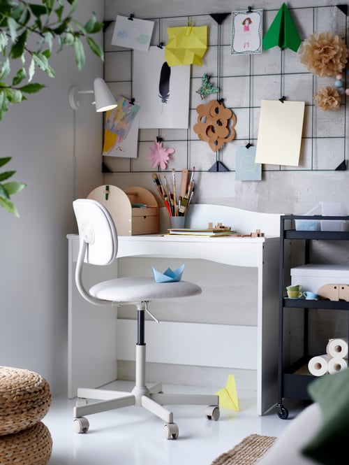 A white SMÅGÖRA children’s desk with a swivel chair, a black trolley, a memo board with clips and drawings, and a wall lamp.