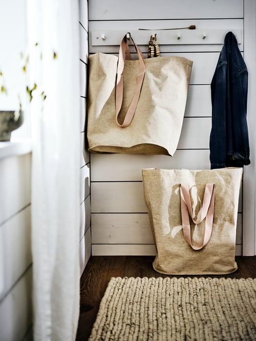 Un mur lambrissé blanc avec une rangée de crochets sur lesquels sont suspendus un grand sac beige et une veste. Un sac beige repose sur le sol.