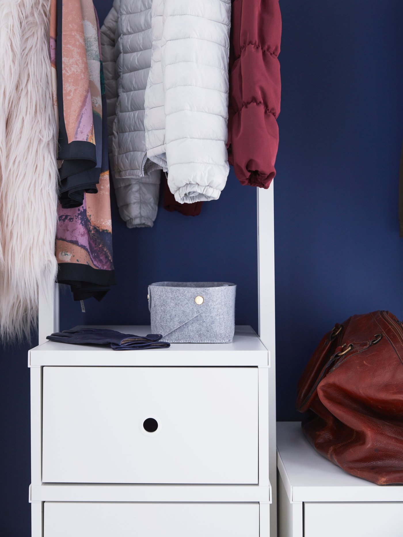 An ELVARLI drawer unit with a small grey storage box on top, with a row of clothing hanging above it against a blue wall.