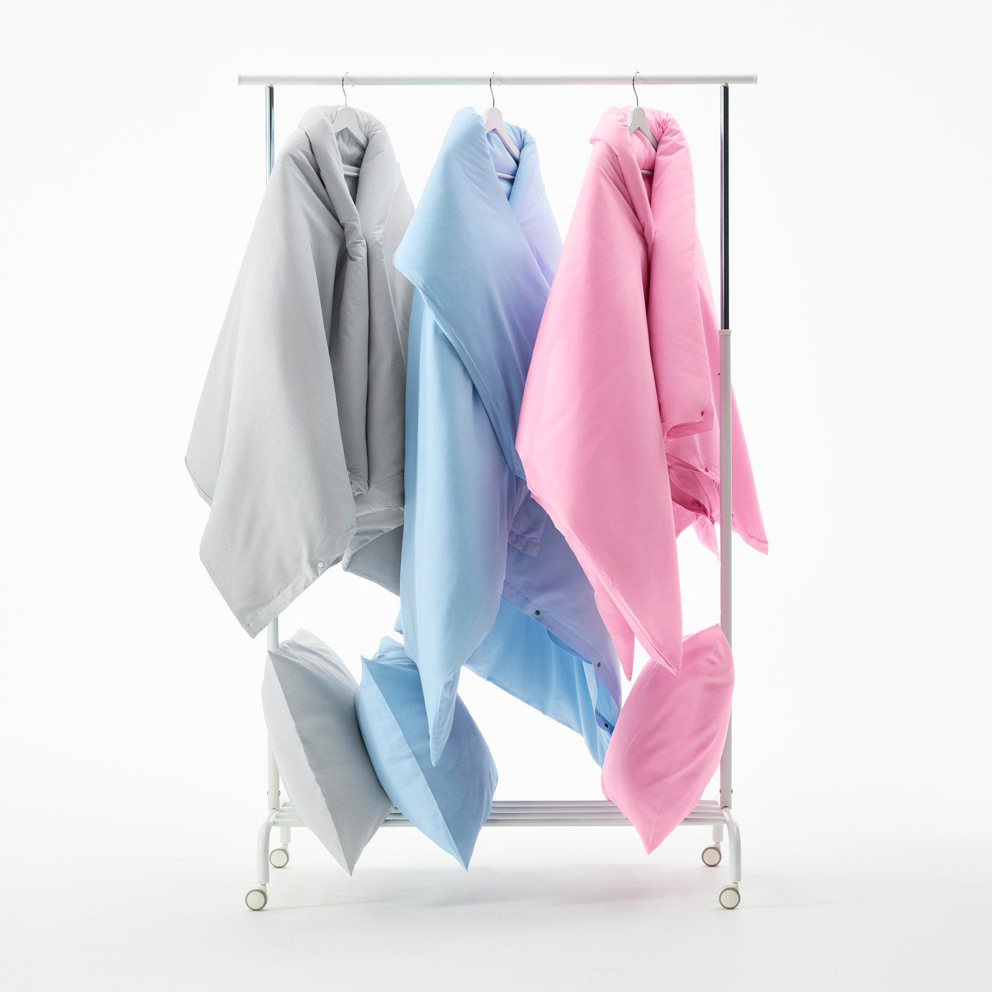 Three PILTANDVINGE duvet sets in grey, light blue and pink hang on a clothing rack. On a shelf below, matching pillows.