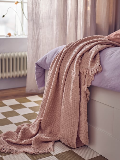 Różowa narzuta HORNMAL położona na łóżku, obok liliowej poszwy na kołdrę i 2 poszewek na poduszki, w nowoczesnej sypialni.
