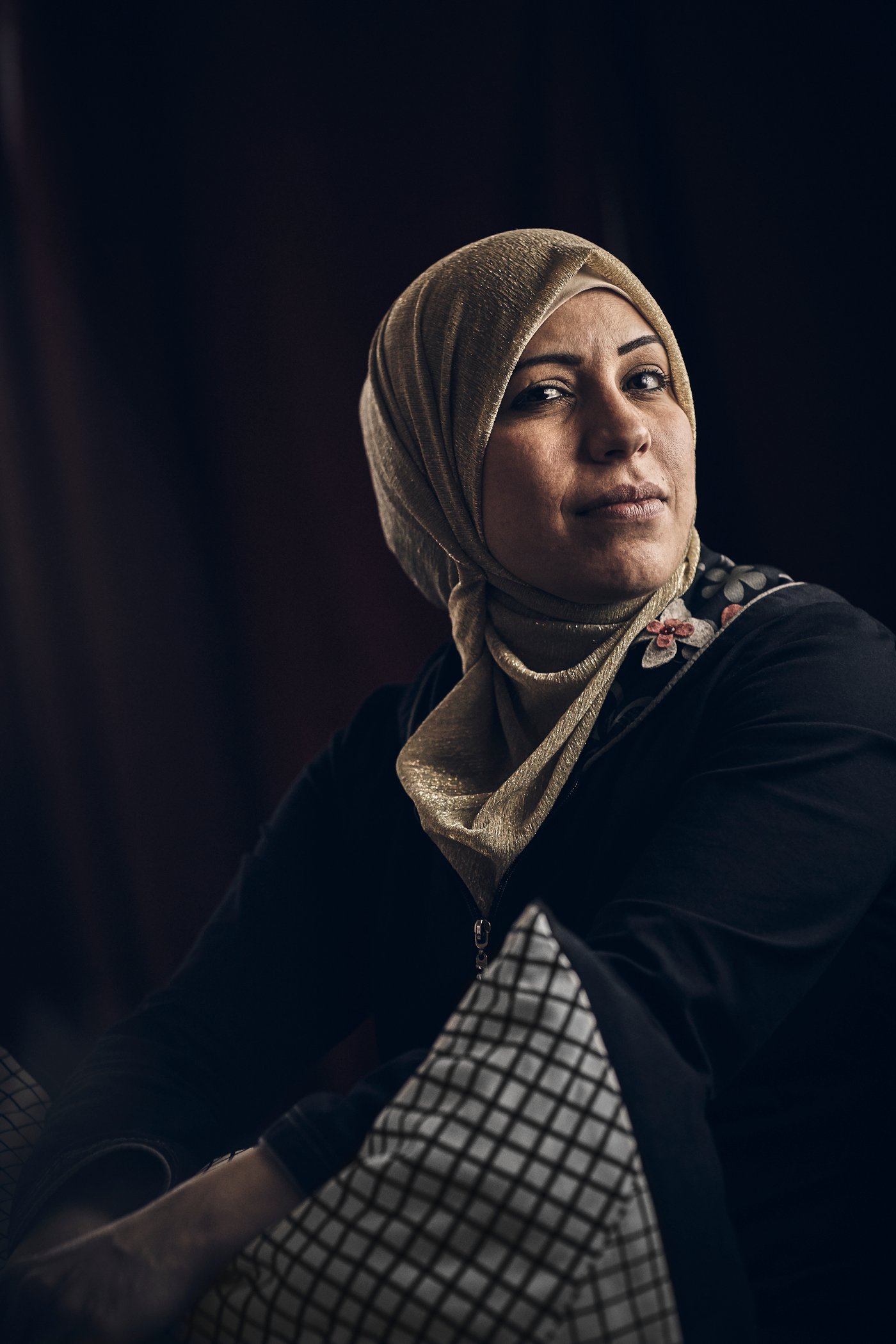 A middle eastern woman wearing a black top with an embroidered collar, and a beige hijab, against a black background.