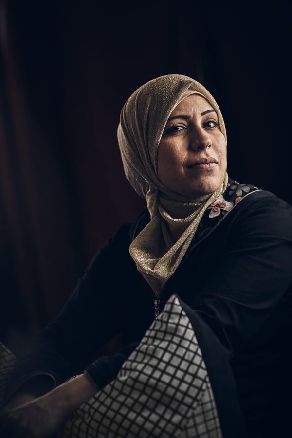 A middle eastern woman wearing a black top with an embroidered collar, and a beige hijab, against a black background.